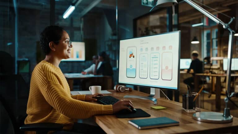 Woman designing mobile app interface on large monitor in modern office.