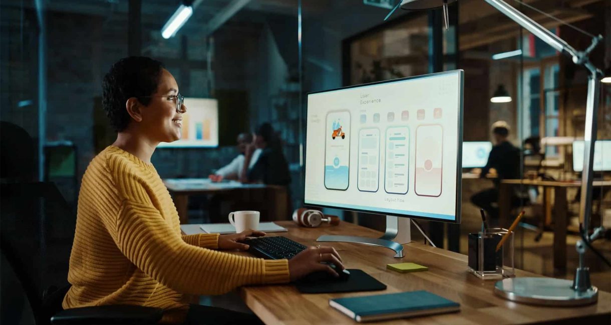 Woman designing mobile app interface on large monitor in modern office.